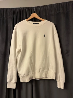 Vit sweatshirt från Polo Ralph Lauren - Vit sweatshirt från Polo Ralph Lauren med klassisk rund hals och broderad logga på bröstet. Tröjan har långa ärmar, ribbade muddar och är tillverkad i mjuk bomull. Perfekt för en clean och stilren look.