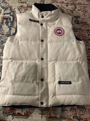 Beige dunväst från Canada Goose - Snygg beige dunväst från Canada Goose med hög krage och två stora fickor framtill. Västen har klassisk logotyp-patch på bröstet och svarta tryckknappar. Perfekt för kalla dagar och har en stilren, quiltad design.
