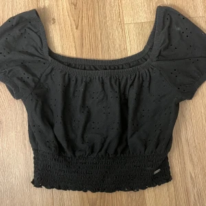 Svart blus från Hollister med puffärm - Supersöt svart blus från Hollister med puffiga korta ärmar och bred smockad midja. Blusen har broderade hålmönster och en rundad, lite djupare halsringning. Perfekt för dig som gillar en feminin och trendig look. Materialet är mjuk bomull.