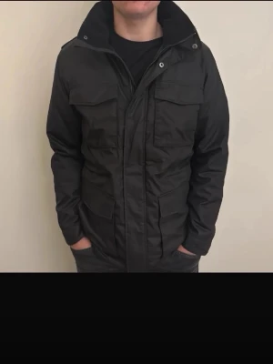 Svart parkajacka med fickor - Svart parkajacka med hög krage och flera stora fickor framtill. Jackan har dragkedja och tryckknappar, samt långärmade ärmar. Materialet är troligtvis polyester och jackan har en normal passform. Perfekt för dig som gillar stilren och funktionell design.