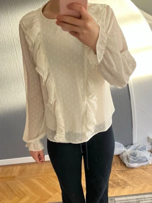Vit blus med volang och små prickar  - Detta är en vit blus från Gina Young med små prickar och volang där fram och vid armarna. Den har ett litet tunt inre tyg så den inte ska vara så genomskinlig. Storlek 158-164 