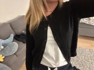 Svart kofta med knappar - En enkel svart kofta med rund hals och knappar framtill. Koftan har långärmade ärmar och en klassisk, normal passform. Perfekt att slänga över en t-shirt eller linne för en chill look. Materialet känns mjukt och bekvämt mot huden.