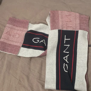 halsduk från GANT - Snygg stickad halsduk från GANT med breda ränder i grått, marinblått och vinrött. GANT-loggan är tydligt broderad i vitt på den mörkblå delen. Perfekt accessoar för att lyfta din vinterstil.