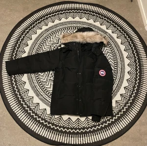 Svart Canada Goose dunjacka med päls - Svart dunjacka från Canada Goose med ikonisk patch på ärmen och fluffig päls runt huvan. Jackan har lång ärm, dragkedja med logotyp och knappslå framtill. Perfekt för kalla vinterdagar och har en clean, klassisk look.