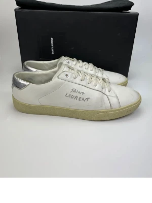 Saint Laurent vita sneakers med silverdetalj - Snygga vita sneakers från Saint Laurent med handskriven logga på sidan och silverfärgad häl. Skorna har rund tå, snörning och platt sula i beige ton. Tillverkade i skinn för en lyxig känsla och stilren look. Perfekta för dig som gillar designer och streetstyle.