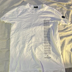 Vit Moncler t-shirt med tryck - ÄKTA. Vit t-shirt från Moncler med svart logotyp-tryck i vertikal rad på framsidan och en svart Moncler-patch på ärmen. Klassisk rund hals och korta ärmar. Tillverkad i mjuk bomull som känns skön mot huden. Perfekt för dig som gillar stilrena designer med tydlig branding.