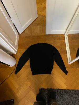 Svart stickad tröja Louis Vuitton - Svart stickad tröja från Louis Vuitton med klassisk rund hals och långa ärmar. Tröjan har en stilren design och en brun Louis Vuitton-logga på ryggen. Perfekt för dig som gillar exklusiva märken och enkel stil. Sitter som en M - modifierad i längden.