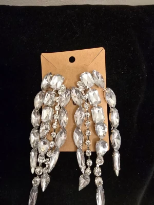 Glittriga silverfärgade örhängen - Säljer ett par statement örhängen med stora, glittriga stenar i silverfärg. Örhängena har flera rader av facetterade kristaller som ger en lyxig och iögonfallande look. Perfekta för att lyfta din outfit till nästa nivå.