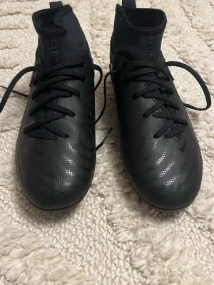 Svarta Nike fotbollsskor strl 36.5 - Säljer ett par svarta Nike fotbollsskor med högt skaft och snörning. Skorna har en stilren design med diskret Nike-logga på sidan och mönstrad ovandel. Materialet är syntet och skon har rund tå samt platt sula för bra grepp på planen.