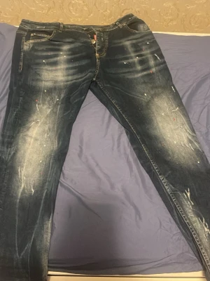 Mörkblå Dsquared2 jeans med färgstänk - Säljer ett par snygga mörkblå jeans från Dsquared2 med coola slitningar och färgstänk. Jeansen har klassisk femficksdesign, tydliga tvättade detaljer och Dsquared2-logga vid gylfen. Perfekt för dig som gillar streetstyle och vill sticka ut. Används it i länge 