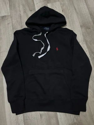 Svart hoodie – Polo Ralph Lauren - Klassisk svart hoodie med den röda Ralph Lauren-loggan på bröstet. Mjuk och skön kvalitet, känguruficka framtill och justerbara vita snören vid huvan.