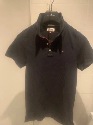 Svart kortärmad skjorta Tommy Jeans - Svart kortärmad skjorta från Tommy Jeans med klassisk krage och två knappar framtill. Liten logotyp på bröstet och diskret röd detalj vid knappslån. Skjortan är gjord i mjuk bomull och har en normal passform som är enkel att styla.