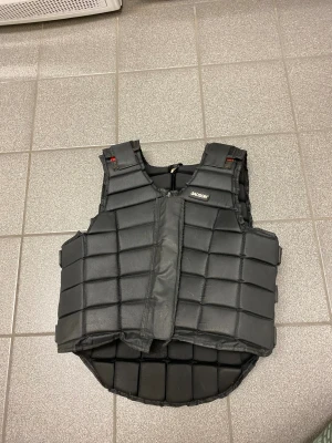 Svart säkerhetsväst från Jacson - Säljer en svart säkerhetsväst från ridsport med quiltad design och justerbara remmar på sidorna som man kan justera. Västen har dragkedja framtill och är tillverkad i slitstark syntet med vadderade paneler för extra skydd. Perfekt för dig som vill ha trygghet vid ridning.