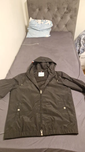 Svart vindjacka från Moncler med luva - Svart vindjacka från Moncler med huva och dragkedja framtill. Jackan har två fickor med tryckknappar, elastiska muddar och diskret Moncler-logga på ärmen. Lätt och smidig modell i syntetmaterial, perfekt för blåsiga dagar.