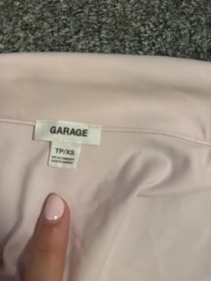 Ljusrosa polotopp från Garage XS - Säljer en ljusrosa polotopp från Garage i storlek XS. Toppen har korta ärmar, klassisk krage och tre knappar framtill. Materialet är mjukt och stretchigt, perfekt för en chill och snygg look. Enkel design utan mönster, passar dig som gillar stilrena plagg.