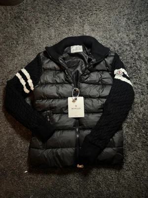 Svart Moncler pufferjacka med stickade ärmar - Svart pufferjacka från Moncler med stickade, kabelmönstrade ärmar och vita ränder på ena armen. Jackan har dragkedja framtill, ribbad krage och muddar. Moncler-logga på ärmen och quiltad framsida i syntetmaterial, den passar som storlek M och en liten L i mans storlek, Snygg och unik design som sticker ut, var inte rädd för att fråga om du har frågor!😁