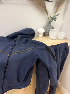Mörkblå Nike hoodie + byxa - Säljer en mörkblå hoodie och byxa från Nike med dragkedja och huva. Tröjan har svarta detaljer, snörning vid huvan och en cool ficka med dragkedja på ärmen. Materialet är mjukt och känns som bomull, perfekt för chill eller träning. Nike-logga på bröstet.