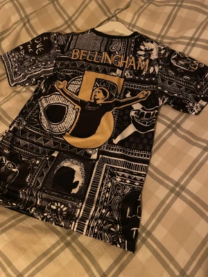 Adidas Real Madrid Bellingham tröja 176 - Säljer en limited edition Real Madrid tröja från Adidas med Bellingham-tryck. Tröjan har svart och guld paisley-mönster, coola grafiska detaljer och guldiga Adidas-ränder på axlarna. Materialet är lätt polyester och perfekt för fotboll eller att rocka på stan.