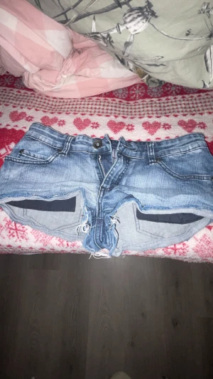 Blå jeansshorts från Denim Jeankmakers - Säljer ett par blå jeansshorts från Denim Jeankmakers med klassisk femficksdesign och råa, fransiga kanter. Shortsen har normal passform, knapp och dragkedja framtill samt snygga slitningar för en chill vibe. Perfekta för varma dagar och enkel att matcha med allt.