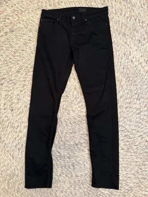 Svarta Tiger of Sweden jeans Evolve - Svarta jeans från Tiger of Sweden, modell Evolve. Slimmad passform med klassisk femficksdesign och svarta nitar. Snyggt minimalistiskt utseende och diskret logga vid fickan. Perfekt för dig som gillar stilrena och smala jeans.