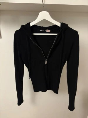 Svart stickad hoodie med dragkedja Gina Tricot - Svart stickad hoodie från Gina Tricot med dragkedja framtill och huva. Tröjan är stickad och har långa ärmar samt ribbad nederkant. Snygg croppad passform som ger en trendig look. Tröjan är i lent material och tajt i passformen😊
