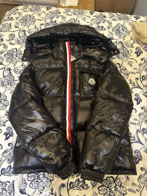 Svart pufferjacka från Moncler - Säljer en svart glansig pufferjacka från Moncler med huva och ikonisk logga på bröstet. Jackan har röd och vit rand längs dragkedjan, quiltad design och innerfoder med serietidningsmönster. Perfekt för kalla dagar och riktigt snygg streetstil.