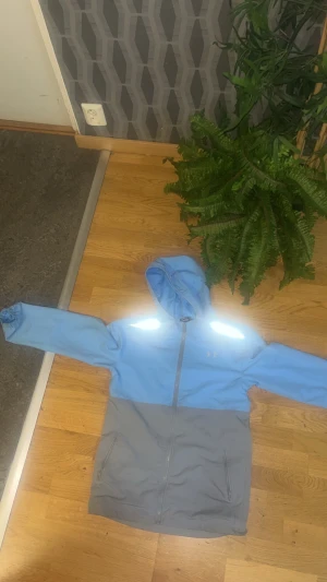 Blå och grå vindjacka från Under Armour - Snygg tvåfärgad vindjacka från Under Armour i ljusblått och grått. Jackan har huva, hel dragkedja framtill och två sidofickor med dragkedja. Reflekterande detaljer på axlarna och elastiska muddar vid ärmslut. Perfekt för dig som gillar sportig stil.
