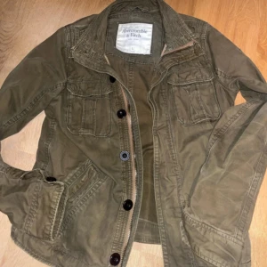 Olivgrön overshirt från Abercrombie & Fitch - Säljer en olivgrön overshirt från Abercrombie & Fitch i storlek L. Jackan har flera fickor framtill, knappar och dragkedja, samt klassisk krage. Perfekt för lager-på-lager och chill stil. Materialet är bomull och passformen är normal.