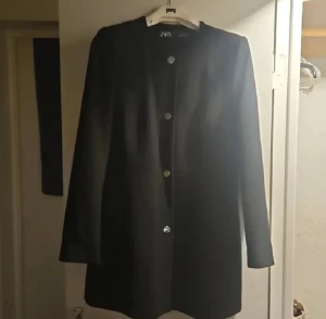 Svart kappa från Zara med knappar - Stilren svart kappa från Zara med rund hals och raka linjer. Jackan har fyra silvriga knappar framtill och långa ärmar. Perfekt för dig som gillar minimalistisk design och vill ha en klassisk look.