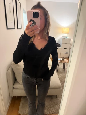 Svart långärmad topp med spets - Svart långärmad topp med v-ringad hals och snygga spetsdetaljer längs urringningen. Modellen är figurnära och har en stilren look som funkar till jeans eller kjol. Materialet känns mjukt och stretchigt, perfekt för dig som gillar en enkel stil
