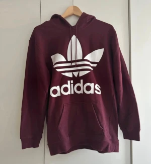 Vinröd Adidas hoodie med logga - Säljer en vinröd hoodie från Adidas med klassiska trefoil-loggan i vitt på framsidan. Tröjan har huva med snörning, magficka och långa ärmar. Perfekt för både vardag och chill dagar