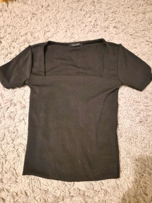 Svart ribbad topp från Amisu XXS - Svart ribbad topp från Amisu i storlek XXS. Toppen har en fyrkantig halsringning och korta ärmar, vilket ger en clean och modern vibe. Materialet är mjukt och stretchigt, perfekt för att styla med dina favoritjeans eller kjol.