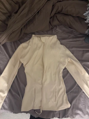 Beige träningsjacka med dragkedja - Säljer en beige, figurnära träningsjacka med hel dragkedja framtill och hög krage. Jackan har långa ärmar och är tillverkad i ett stretchigt syntetmaterial som passar perfekt till gymmet. Snygg och enkel design som är lätt att matcha med träningskläder.