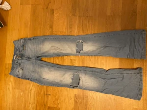 Bootcut jeans med slitningar från Nelly - Snygga ljusblå bootcut jeans från Nelly i storlek 34. Jeansen har slitna detaljer på knäna, klassisk femficksdesign och dubbla knappar i midjan. Tillverkade i mjukt denim med gul kontrastsöm och lätt tvättad look.