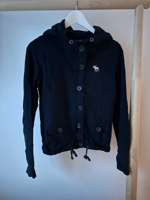 Abercrombie & Fitch hoodie – mörkblå - Säljer en mörkblå hoodie i avslappnad, sportig stil. Jackan har knäppning framtill, två fickor med knappar, dragsko längst ner och huva. Broderad logga på bröstet. Kids L/XL men passar som vuxen S.