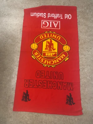 Manchester United röd frotté badhandduk - Stor röd badhandduk med Manchester Uniteds klubbmärke i gult och svart, samt texten Old Trafford Stadium och AIG. Handduken har svart tryck med klubbnamnet och siluetter i hörnen. Perfekt för fotbollsfans som vill visa sitt lag på stranden eller gymmet.