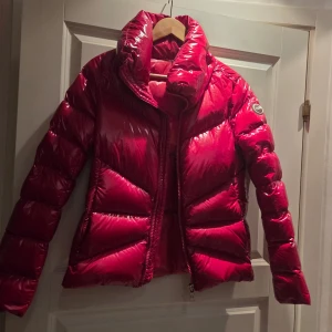 Röd jacka från Colmar - Säljer en riktigt snygg röd pufferjacka från Colmar Originals med glansig finish och hög krage. Jackan har en tydlig logga på ärmen, dragkedja framtill och är vadderad för extra värme. Perfekt för dig som vill sticka ut i vinter och hålla dig varm.