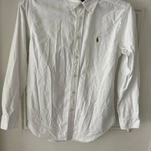 Vit skjorta från Ralph Lauren - Klassisk vit skjorta från Ralph Lauren med broderad logga på bröstet. Skjortan har lång ärm, knappar framtill och krage. Perfekt till både avslappnade och mer uppklädda tillfällen. Modellen är 173 och väger 60 kg