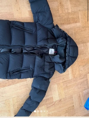 Svart Moncler dunjacka med huva - Säljer en svart dunjacka från Moncler med huva och dragkedja. Jackan har två snedställda fickor med dragkedja och en klassisk puffer-design. Finns en liten repa på baksidan av rackan men som är lagad hos skreddare (bild3).Luvan är avtagbar. Har även lagt ut jackan på vinted där det finns äktighetsbevis samt fler bilder, kan även skicka detta vid önskan. Perfekt för kalla vinterdagar