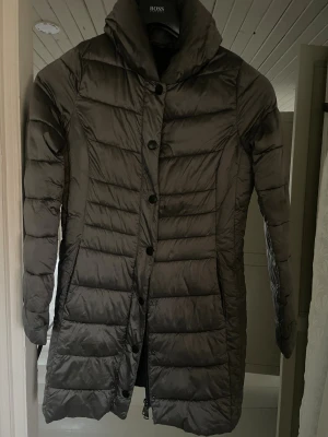 Mörkgrå dunjacka från BOSS - Snygg mörkgrå dunjacka från BOSS med hög krage och långa ärmar. Jackan har knappar framtill och är quiltad för extra värme. Perfekt för kyliga dagar och har en stilren, lång passform som ger en modern look.