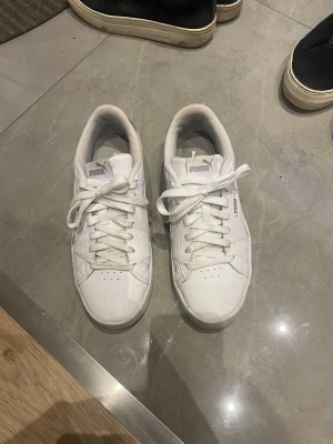 Vita Puma sneakers med silverdetalj - Fräscha vita Puma sneakers med klassisk design och silverdetalj på hälen. Skorna har snörning, platt sula och rund tå. Materialet är skinn och syntet, vilket gör dem både snygga och bekväma. Perfekta för dig som gillar stilrena sneakers med en liten twist.