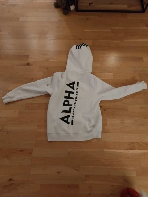 ALPHA INDUSTRIES HOODIE, VIT  - En Alpha industries hoodie med svart text på ryggen. Passform medium. 