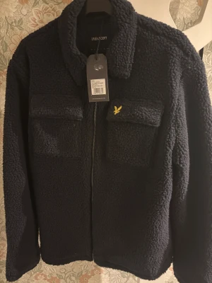 Mörkblå teddyjacka från Lyle & Scott - Mysig mörkblå teddyjacka från Lyle & Scott med två bröstfickor och dragkedja framtill. Jackan har klassisk krage och den ikoniska gula loggan broderad på ena fickan. Perfekt för dig som vill ha en stilren och varm look.