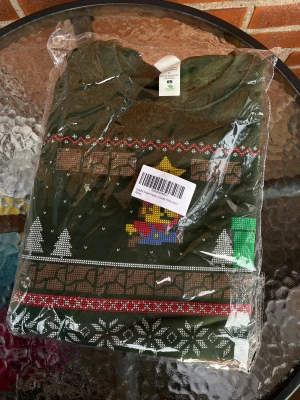 Ny Grön jultröja tröja med Super Mario motiv. Strl L - Säljer en mörkgrön jultröja från Graphic Impact med Super Mario-motiv och klassiska retrofigurer. Tröjan har lång ärm, rund hals och tryck med snöflingor, stjärnor och spelikoner i rött, gult, blått och vitt. Perfekt för dig som älskar gaming och coola prints.