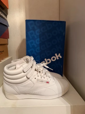 Reebok classic FREESTYLE HI HIGH-TOP TRAINERS - Höga sneakers - Snygga vita Reebok sneakers med högt skaft och dubbla kardborreband upptill. Skorna har snörning, rund tå och platt sula. Tillverkade i skinn med mjuk vaddering runt ankeln för extra komfort. Klassisk Reebok-logga på sidan.