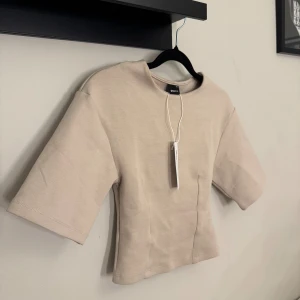 Beige fitted topp från Gina Tricot - Säljer en fitted topp från Gina Tricot i färgen chateau gray. Toppen har korta ärmar, rund halsringning och en croppad passform. Tillverkad i mjuk bomullsmix som känns skön mot huden. Perfekt att styla med jeans eller kjol.