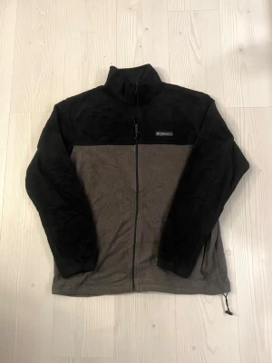 Svart och grå fleecejacka Columbia - Mjuk och skön fleecejacka från Columbia i storlek L. Jackan är tvåfärgad med svart upptill och grå nedtill, har hel dragkedja framtill och hög krage. Perfekt för kyliga dagar och outdoor-häng. Diskret logga på bröstet och justerbar nederkant med snörning.