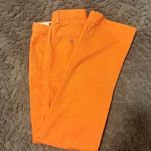 Orange chinos från Polo Ralph Lauren - Snygga orange chinos från Polo Ralph Lauren i storlek 30/32. Byxorna är tillverkade i 100% bomull och har klassisk rak passform med bälteshällor och knappstängning. Perfekt för dig som vill sticka ut med färg och stil. Kan bli riktigt snyggt till sommaren om man stylar de bra