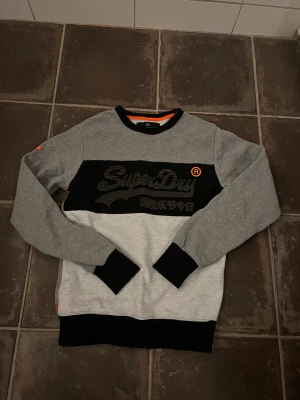 Superdry sweatshirt - Snygg grå och svart sweatshirt från Superdry med stort logotryck på bröstet och japanska tecken. Tröjan har svarta muddar vid ärmslut, hals och nederkant samt orange detalj på ärmen. Perfekt för dig som gillar streetwear och vill ha en cool look.