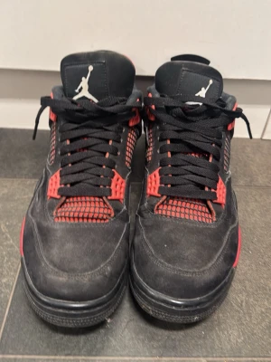 Nike Air Jordan 4 svart/röd sneakers - Nike Air Jordan 4 sneakers i svart och rött med klassisk Jumpman-logga på plösen och hälen. Skorna har meshpaneler, snörning och en platt sula. Insidan är fodrad i rött tyg och detaljerna är i syntet och mocka. Perfekt för dig som gillar streetwear och ikoniska sneakers. Finns box och pris lapp som följer med vid köpet skriv vid fundering
